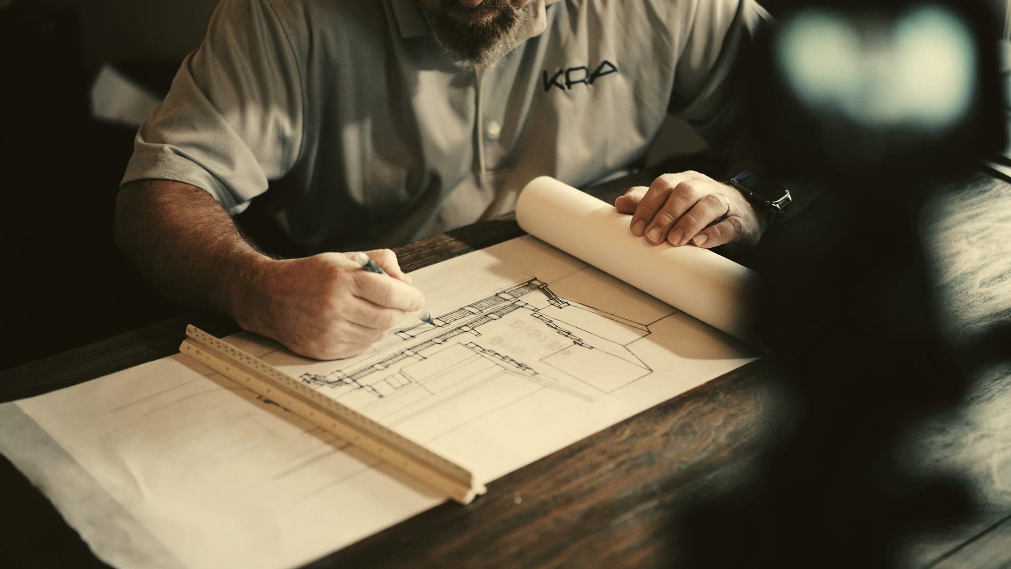 Hands over a rolled technical drawing on a dark bench, a rule laid across it